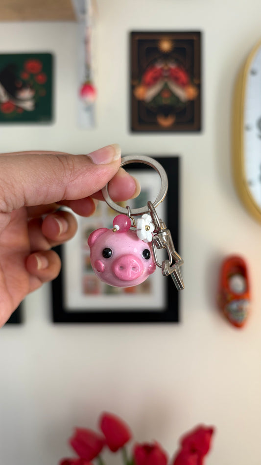 Piggy polymer clay keychain, in-hand scale showing round pink pig face charm with daisy accent and silver clasp