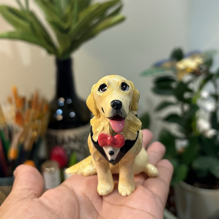 A custom handmade clay sculpture of a golden retriever wearing a red bandana, held in a hand with plants behind