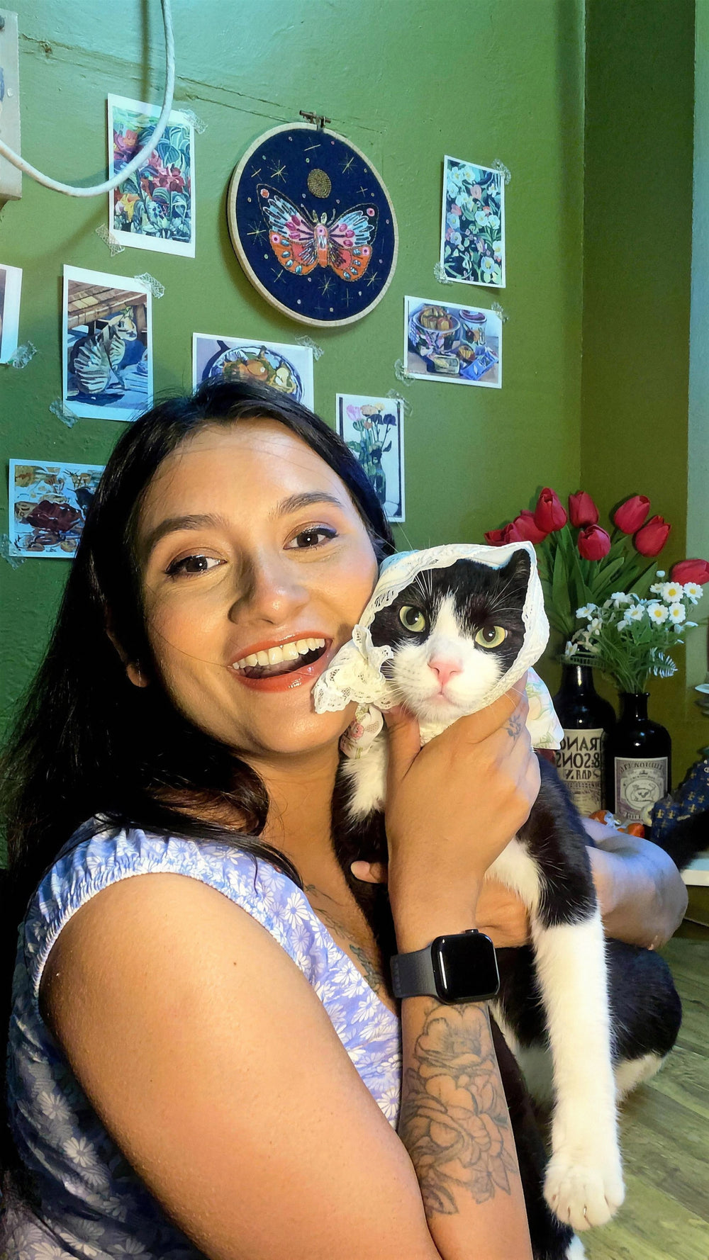 Amba, the maker behind Moon Kiln, smiling with her tuxedo cat in a lace bonnet in the studio, art prints and tulips on the green wall behind them