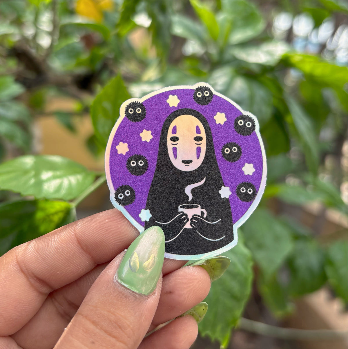 Masked Spirit holographic vinyl sticker, illustrated black-robed spirit with white oval mask holding tea cup on purple background, held in hand