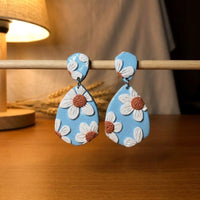 Daisy Blue handmade polymer-clay earrings, displayed on a stand in warm evening light