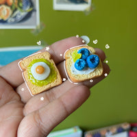Blueberry ricotta toast and avocado egg toast polymer clay keychains side by side, in-hand scale showing miniature food detail